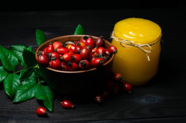 Stüdyoda yeşil dallı kırmızı gül kalçaları ve koyu ahşap arka plandaki kavanozda bal. Tıbbi bakım ve sağlıklı gıda konsepti.