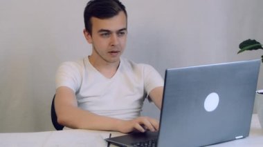 Yoğunlaşmış genç serbest çalışan dizüstü bilgisayara bakıyor, uzaktan evde çalışıyor. Kafa resmi odaklı gülümseyen adam sosyal ağda sohbet ediyor, mesaj yazıyor, internet sörfü yapıyor..