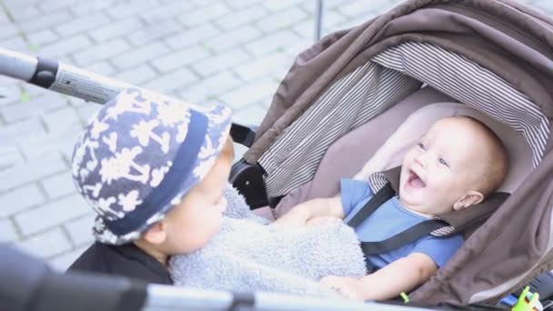青い帽子で男の子を笑っている子供たちは笑顔の幼児馬車のベビーカーを気にします — ストック動画