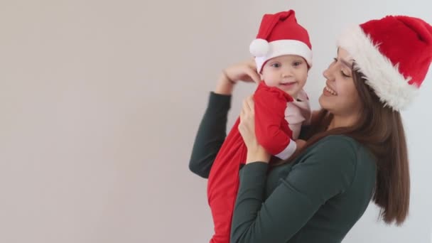 笑顔Caucasianクリスマスファミリーの若いですブルネット長い髪の母で緑のシャツを保持で手かわいい幼児赤ちゃん9ヶ月でサンタクロース赤い衣装 ヘルパー帽子一緒に見るカメラ大好き — ストック動画