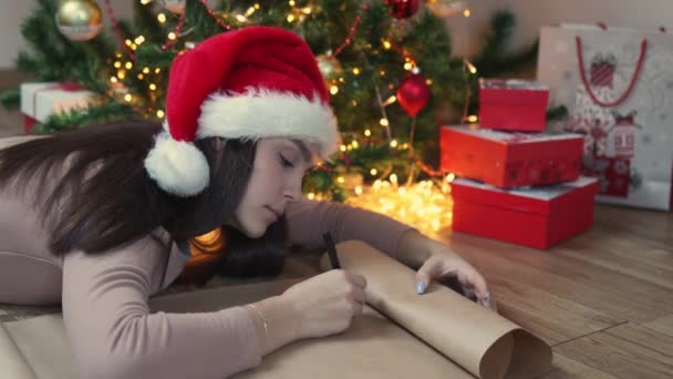 Morena mujer en rojo Santa sombrero escribe carta árbol de Navidad luces cajas de regalo — Vídeos de Stock