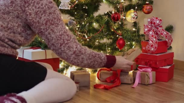 Mano colocando cajas de regalo de artesanía roja bajo luces parpadeantes decoradas árbol de Navidad — Vídeos de Stock