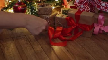 Noel ağacının altında kahverengi el işi kağıdına kırmızı kurdele saran eller.