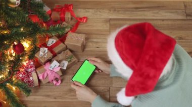 Kadın kırmızı Noel şapkalı akıllı telefon köknar ağacının altında yeşil ekran hediye kutuları veriyor