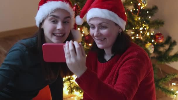 Dos hembras en sombrero de santa verde con teléfono inteligente en el fondo del árbol de Navidad — Vídeos de Stock