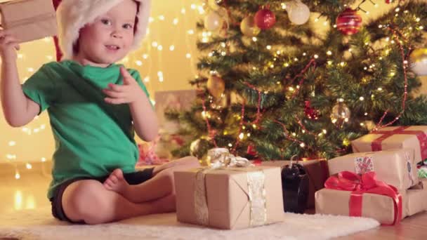 小男孩绿色的衬衫，红红的圣诞老人帽和礼物在枞树下 — 图库视频影像