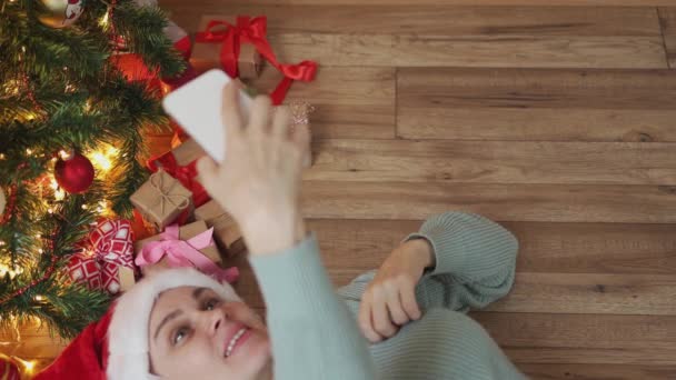 Kvindelig rød julemænd hat liggende på gulvet gran træ gave med videoopkald smartphone – Stock-video