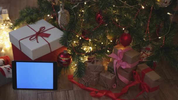 Karácsony piros kézműves ajándékdobozok és tabletta kék mockup képernyő alatt fenyő fa — Stock videók