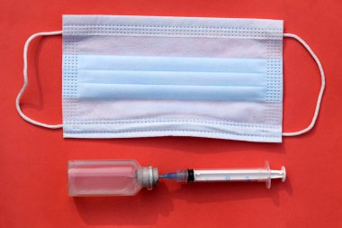 Syringe with vaccine bottle on surgical mask in red background. Healthcare medicine with equipment. Vaccination related objects on red backdrop.