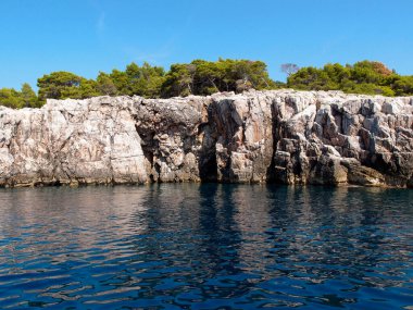 Adanın kayalık kıyılarının güzel manzarası. Arkasında orman ve mavi gökyüzü var. Denizden bir manzara.