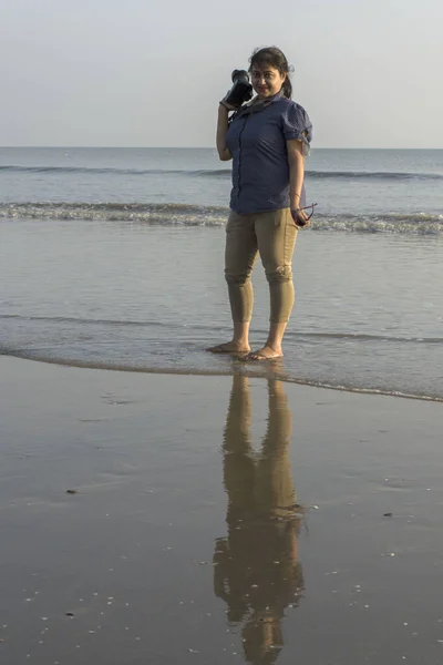 A female model wearing western outfit with camera in hand standing on a sea beach with reflection. Selective focus.