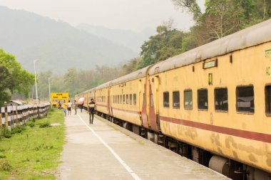 5 Nisan 2022, Shevok, Batı Bengal, Hindistan: Hint Demiryolu 'ndan sarı bir tren Shevok İstasyonu' na vardı ve birkaç yolcu platforma yürüyordu. Arkaplanda seçici odaklı dağlar.