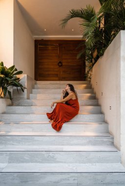 Uzun terasotta elbiseli güzel İspanyol kadın bir binanın girişinde oturmuş gülüyor.