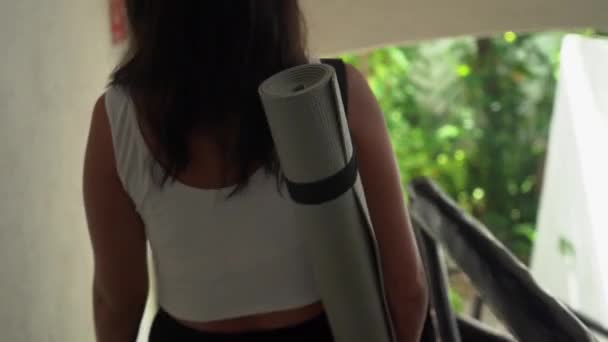 Ung spansktalende kvinde går ned ad trappen med yogamåtte på skulderen – Stock-video