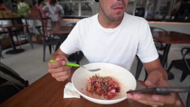 Jonge Spaanse man scrollt op zijn telefoon tijdens het eten van salade in een restaurant — Stockvideo