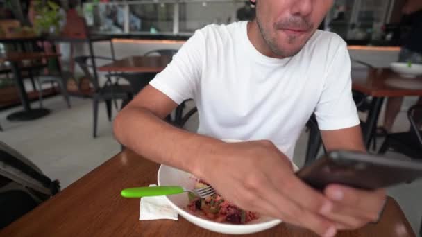 Hispanic young man writes a message on his smart phone while eating salad — Stock Video