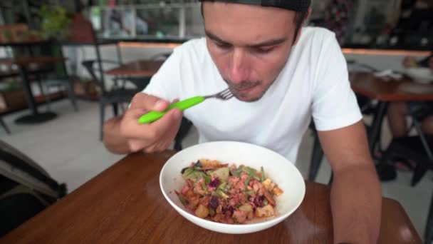Jonge Spaanse man met pet eet salade in een restaurant — Stockvideo