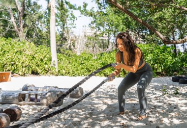Genç İspanyol kadın plaj eko salonunda halatlarla kol egzersizi yapıyor.