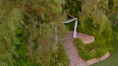 Havadan çekilmiş düğün görüntüleri. Kemerle süslenmiş, dışarıda çiçekler var. Güneşli bir günde parkta düğün düzenleniyor.