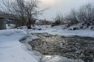 Güneşli bir kış gününde hızlı akan buzsuz bir nehir. Buz, kar ve çalılarla kaplı kıyılar resmi olarak nehir yatağının sınırını çiziyor. Güneşin yansıması. Uzakta köprü var. Rusya, Ural 