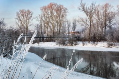 Dondurulmamış suyla nehir manzarası, asma köprü, kar yığınları, kırağı ve güneşli bir kış gününde kıyılarda güçlü kavak ağaçları. Şiddetli dondan dolayı bulanıklık. Bulutlu açık mavi gökyüzü. Rusya, Ural