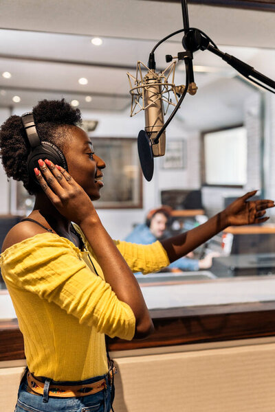 Stock photo of beautiful black woman with headphones using microphone in music studio.