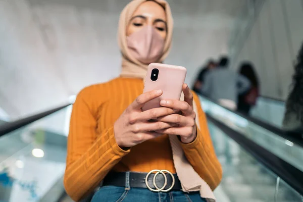美しいですイスラム教徒女性身に着けていますピンクヒジャーブと顔マスクタイプで彼女の携帯電話と使用してエスカレーター — ストック写真