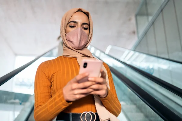 美しいですイスラム教徒女性身に着けていますピンクヒジャーブと顔マスクタイプで彼女の携帯電話と使用してエスカレーター — ストック写真