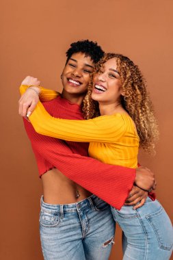Stock photo of cheerful afro women hugging each other in studio shot against brown background.
