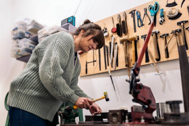 Mücevher atölyesinde çalışan ve çekiç kullanan konsantre bir kadın..