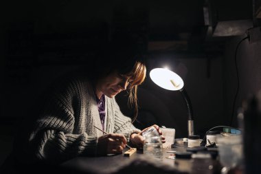 Stock photo of concentrated woman using paint in jewelry workshop.