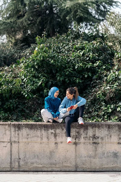 Cheerful muslim woman wearing hijab and sports clothes sitting in the street with her friend and using mobile phone.