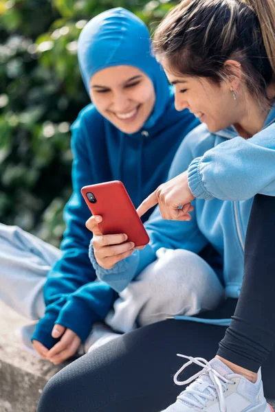Cheerful muslim woman wearing hijab and sports clothes sitting in the street with her friend and using mobile phone.