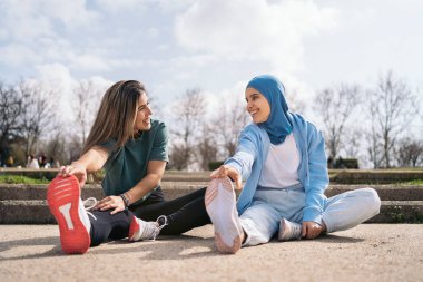 Multicultural female friends sitting in the park stretching their legs after work out. They are wearing sports clothes.