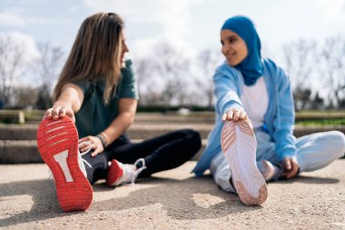 Multicultural female friends sitting in the park stretching their legs after work out. They are wearing sports clothes.