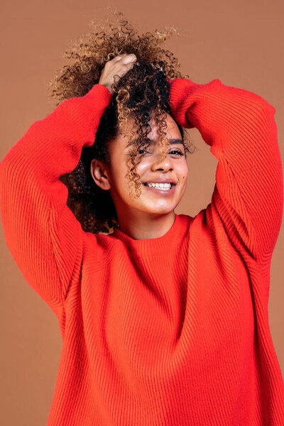 Happy young black woman playing with her curly hair and looking at camera in studio shot against brown background.