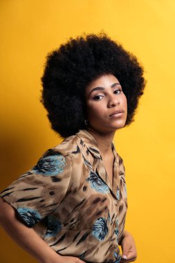 Serious afro woman wearing colorful shirt posing and looking at camera in studio shot against yellow background.