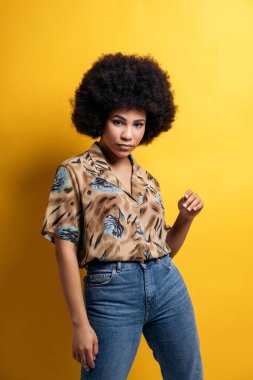 Serious afro woman wearing colorful shirt posing and looking at camera in studio shot against yellow background.