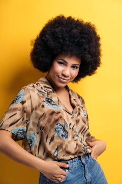 Smiley afro woman wearing colorful shirt smiling and looking at camera in studio shot against yellow background.