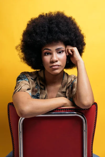 Beautiful afro woman sitting in chair touching her face and looking at camera in studio shot.