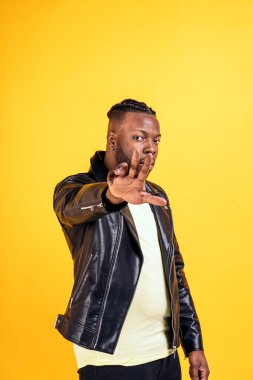 Confident black man wearing black leather jacket posing in studio shot and looking at camera against yellow background.