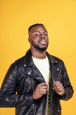 Strong black man wearing black leather jacket posing in studio shot against yellow background.