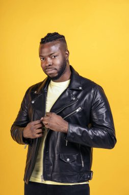 Strong black man wearing black leather jacket posing in studio shot against yellow background.