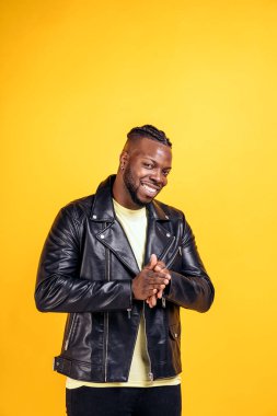 Confident black man wearing black leather jacket smiling in studio shot and looking at camera against yellow background.