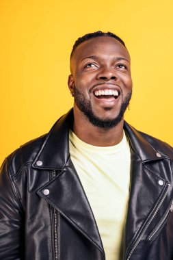 Happy black man wearing black leather jacket laughing in studio shot against yellow background.