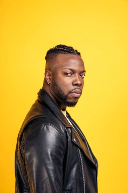 African american man with cool hairstyle and wearing black leather jacket posing in studio shot against yellow background.