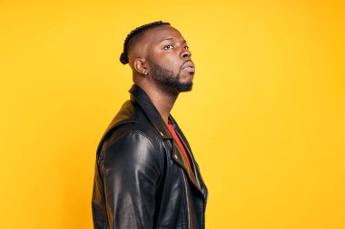 African american man with cool hairstyle and wearing black leather jacket posing in studio shot against yellow background.