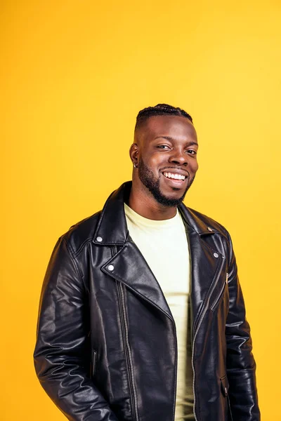 Confident black man wearing black leather jacket smiling in studio shot and looking at camera against yellow background.