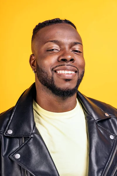 Confident black man wearing black leather jacket smiling in studio shot and looking at camera against yellow background.