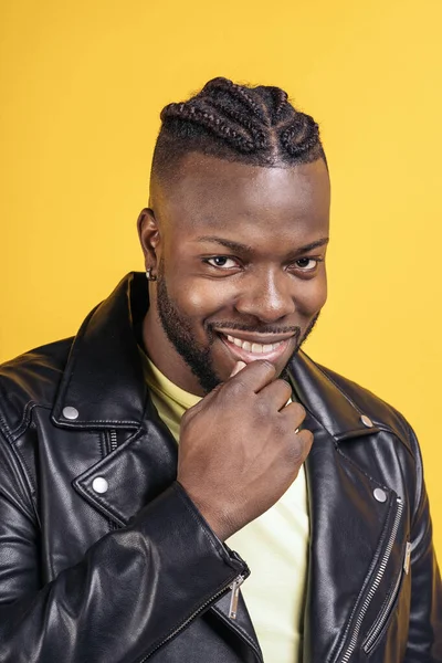 Confident black man wearing black leather jacket posing in studio shot and looking at camera against yellow background.